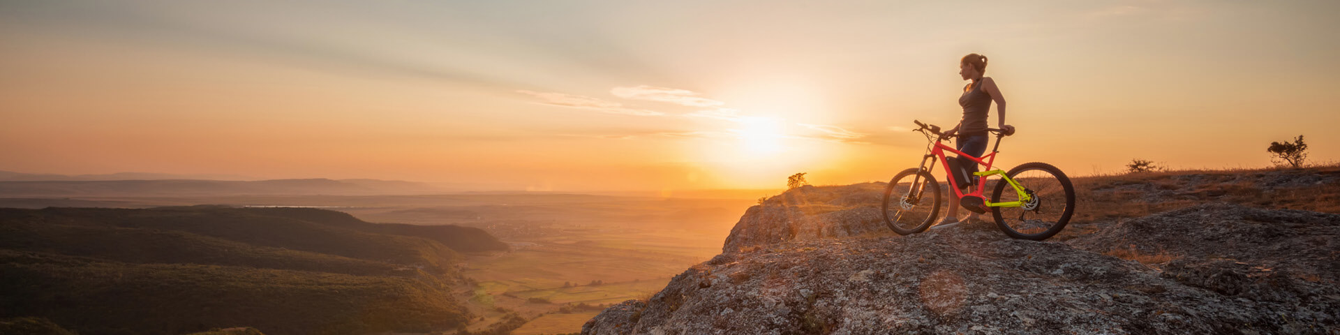 Eine Frau steht mit ihrem Fahrrad auf einem Felsen und betrachtet den Sonnenuntergang